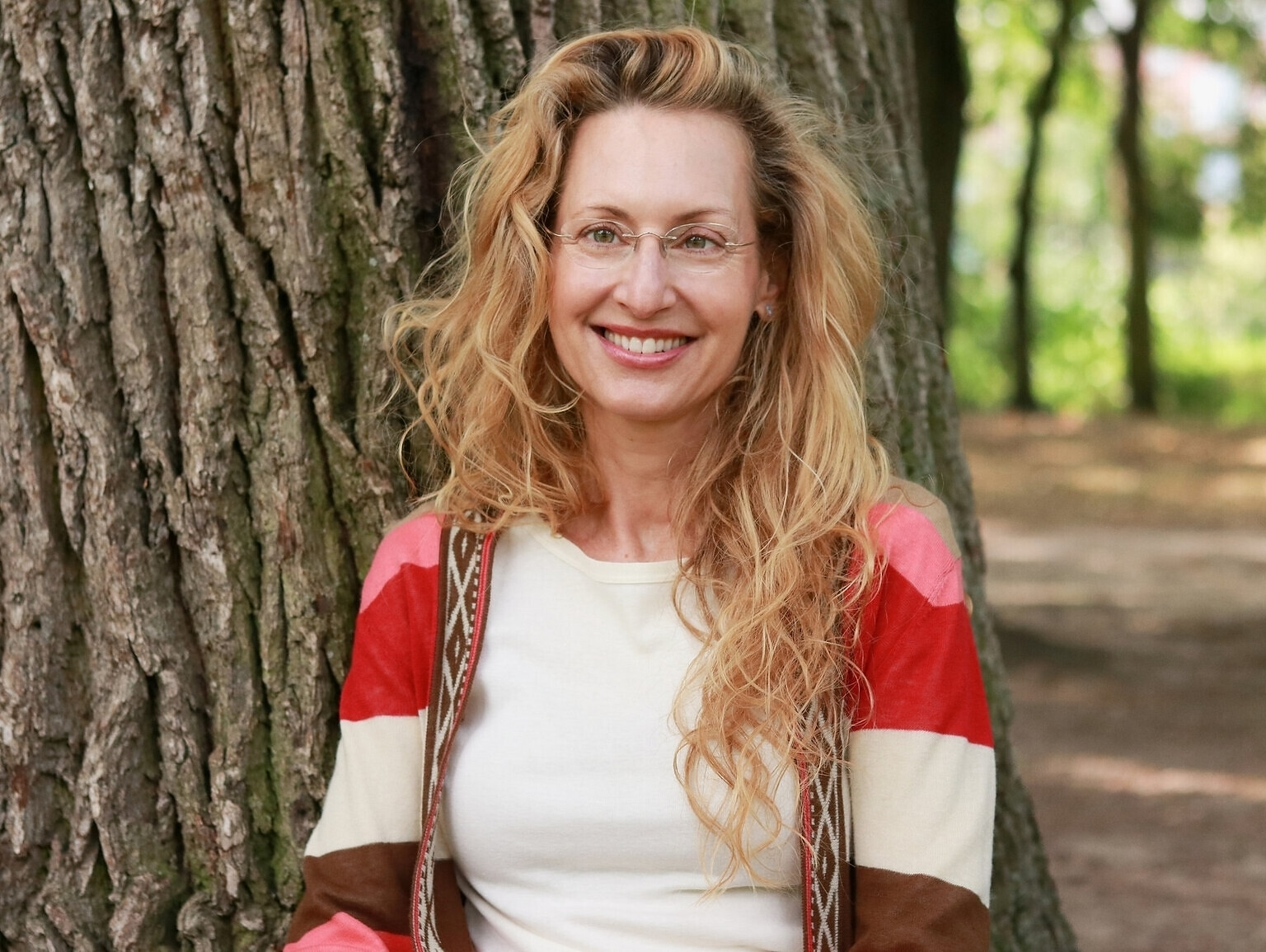Uta Susanne Lowack lächelnd an einen Baumstamm gelehnt, im Hintergrund ein sonniger Park